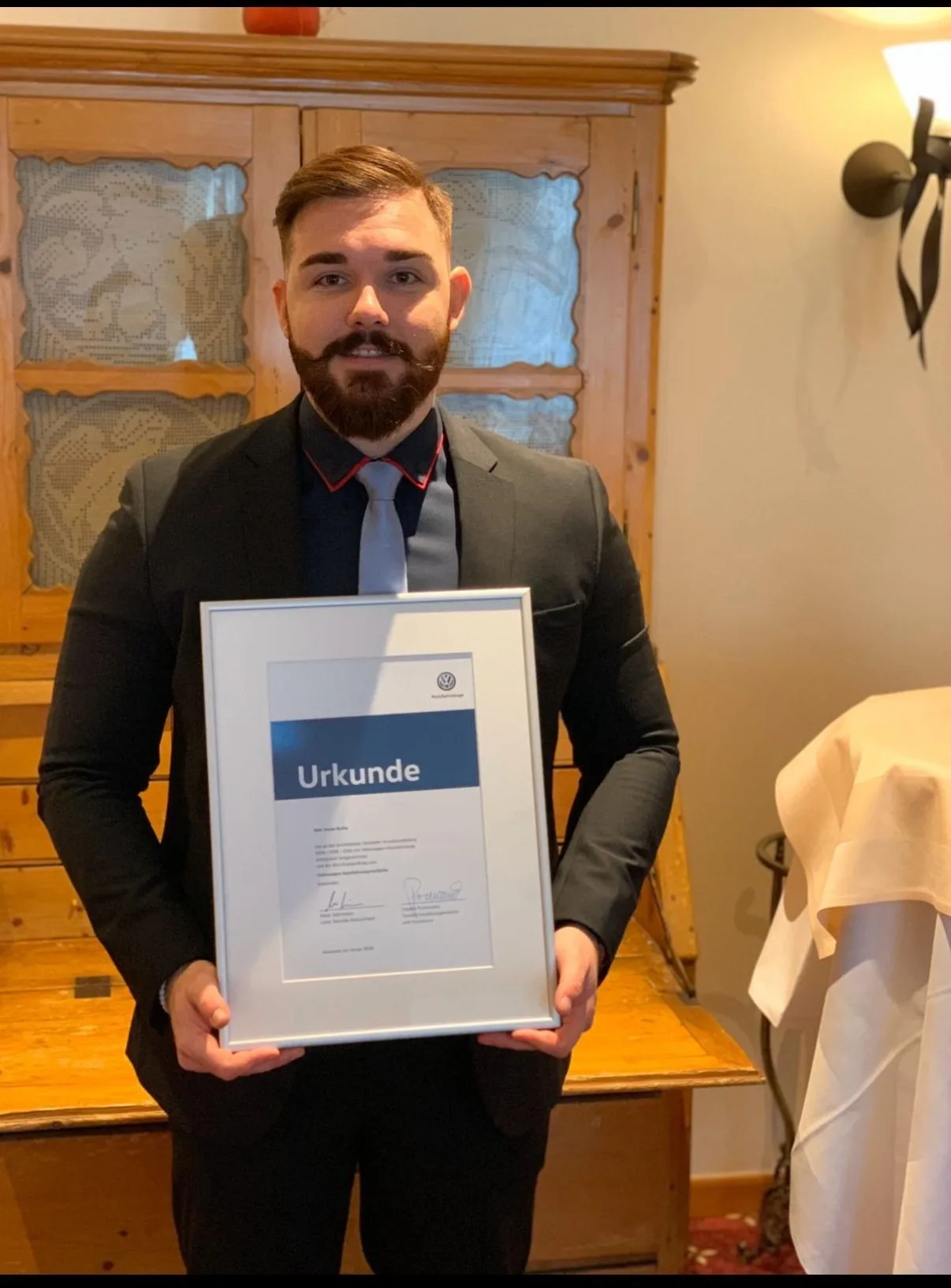 Jonas Huhle im Anzug hält eine gerahmte Volkswagen-Urkunde aus seiner Zeit als Automobilverkäufer.